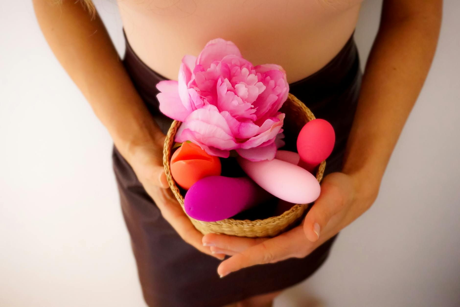 A collection of colorful vibrators in a woven basket with fresh flowers, representing pleasure tools for wellness